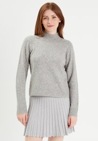 Pull en maille gris clair avec un col montant, des manches longues et un ourlet côtelé. Porté par-dessus une jupe plissée légère, mettant en valeur une texture superposée.
