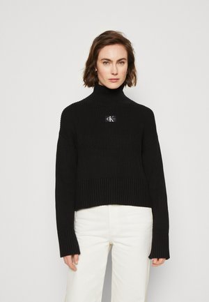 Maglione a collo alto nero a coste, di lunghezza cropped, con un grande logo sul petto e maniche larghe, abbinato a pantaloni bianchi.