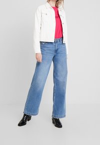 Chaqueta de mezclilla blanca sobre una camiseta roja, jeans de pierna ancha azul claro y botines negros de tacón cuadrado. Tejidos suaves con un diseño casual.