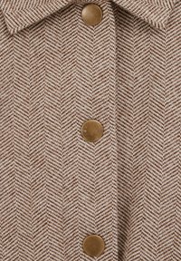Rudas herringbone audinys su tekstūruotais audiniais, centre puikuojasi dideli bronziniai mygtukai. Apykaklės dizainas suteikia struktūrą.