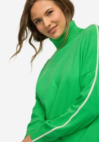 Maglione verde a maglia con collo alto a costine e righe bianche a contrasto lungo le maniche. Tessuto morbido con una vestibilità rilassata.