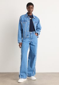 Denimjacke mit ausgefranstem Saum, kombiniert mit hoch taillierten, weit geschnittenen Jeans. Beides in Hellblau, ergänzt durch weiße Sneakers und ein dunkelblaues Hemd.