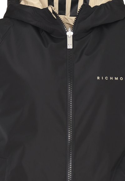 Giacca nera con zip e cappuccio, dotata di una fodera interna beige a contrasto, chiusura frontale con cerniera e logo "RICHMOND" in oro. Texture liscia.