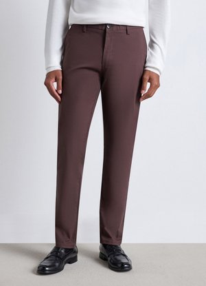 Persona che indossa pantaloni marrone scuro a gamba dritta abbinati a mocassini in pelle nera e top bianco a maniche lunghe su sfondo uniforme.