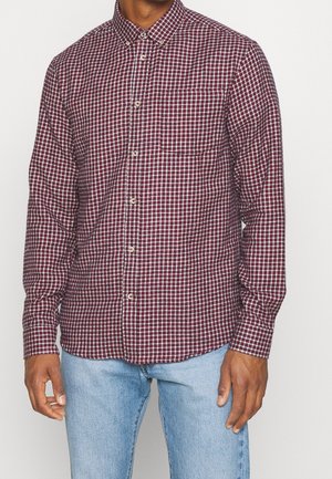 Homme portant une chemise à manches longues boutonnée à carreaux bordeaux et blancs avec une poche poitrine, et un jean bleu clair, posant devant un fond uni.