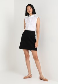 Camicia bianca a maniche corte con colletto abbinata a una gonna nera a trapezio. Il modello indossa delle ballerine beige, in piedi su uno sfondo chiaro.