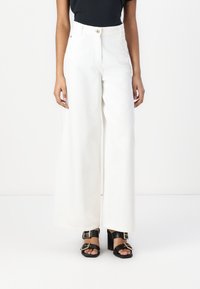 Pantalones anchos blancos de mezclilla, con una cintura alta, cierre de botón en la parte frontal y una textura suave. Combinados con sandalias de tacón negras.