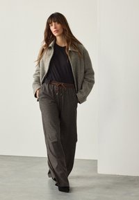 Gray textured jacket, black t-shirt, patterned dark trousers with brown drawstring. Model stands against a neutral background.