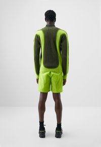 Roheline kudumiga kampsun tekstuurse disainiga, koos erksate laimiroheliste lühikeste pükstega. Kandmine mustade sokkide ja kingadega, vaade tagant.