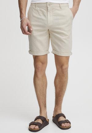 Beige, rolled-up shorts made of lightweight material, featuring front pockets and a button closure. Worn with brown sandals.
