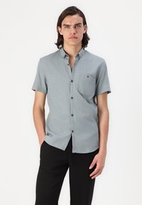 Camisa de botones de manga corta, de color azul claro y fabricada con un tejido texturizado, que presenta un bolsillo en el pecho y botones oscuros. Combinada con pantalones negros.