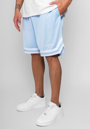 Man draagt lichtblauwe basketbalshorts met witte strepen en witte sneakers, staand met één hand in de zak tegen een grijze achtergrond.