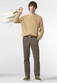 Un pull en tricot beige avec une texture côtelée, un pantalon chino marron, des baskets en daim vert et une ceinture à motifs. Un sac fourre-tout blanc est tenu dans une main.