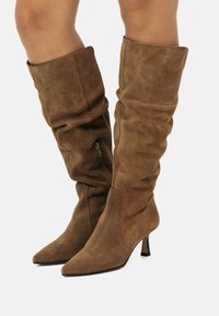 Bottes en daim marron jusqu'au genou avec un design flou, un bout pointu et un talon fin. Équipées d'une fermeture éclair latérale pour un enfilage facile.