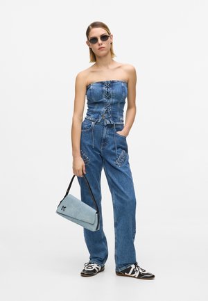 Kobieta w bezramkowym dżinsowym topie typu gorset i dopasowanych dżinsach ze sznurowanymi detalami, trzyma jasnoniebieską torebkę, nosi czarno-białe buty i okulary przeciwsłoneczne.