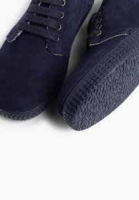 Zapatos de ante azul marino con suela de goma texturizada, que cuentan con cordones redondos y ojales de metal, destacando el diseño elegante y el material del zapato.
