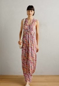 Robe longue sans manches avec un motif zigzag multicolore vibrant, en tissu léger et avec un décolleté en V. Le modèle porte un petit sac.