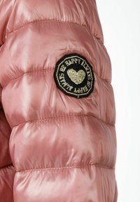 Rosa puffjacka med en glänsande yta, med en rund svart lapptäck med broderad text och ett hjärtmönster på ärmen.