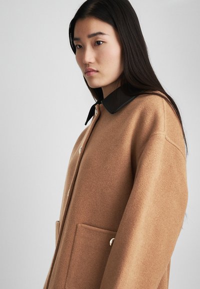 Cappotto di lana marrone con collo in pelle nera, grandi tasche frontali e bottoni in metallo argentato. Design rilassato e texture morbida.