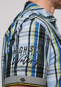 Camicia a maniche corte a quadri nei colori blu, verde e nero. Presenta testo ricamato, rifiniture a contrasto e un dettaglio con bottone sulla manica.