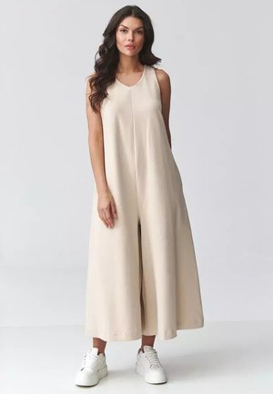 Kobieta z długimi ciemnymi włosami, ubrana w bezrękawnik i szerokie beżowe spodnie kombinezonu oraz białe trampki, stoi na tle jednolitego jasnoszarego tła.