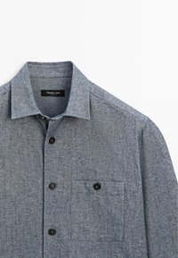 Grijze button-up shirt gemaakt van textuur stof. Heeft een puntige kraag, zwarte knopen en een borstzak aan de linkerkant.