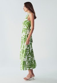 Maxi haljina od bijelog materijala s velikim zelenim uzorcima lišća, bez rukava, strukirana u struku, lepršava suknja, uparena s bijelim klompkama.