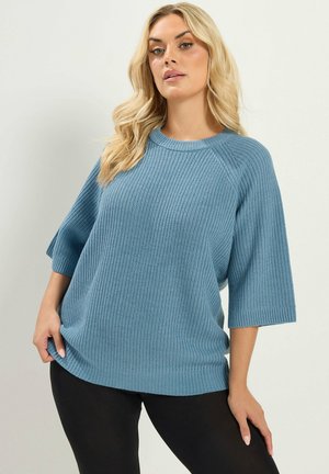 Kvinde med langt blond hår iført en løs blå striktrøje med albuelange ærmer og sorte bukser mod en enkel lys baggrund.