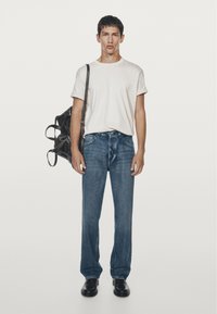 Maglietta bianca, jeans di denim blu, scarpe di pelle nere e una borsa nera con tracolla. Il modello è in piedi davanti a uno sfondo semplice.