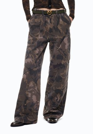 Persona che indossa pantaloni ampi mimetici marroni e neri con grandi tasche, una cintura nera e dorata e scarpe nere, mani nelle tasche.