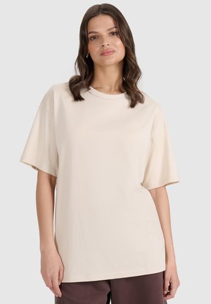 Kobieta o falowanych brązowych włosach do ramion, ubrana w luźny beżowy t-shirt z krótkim rękawem i ciemne spodnie, stojąca na jasnoszarym tle.