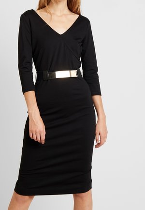 Mujer con un vestido negro hasta la rodilla, con mangas tres cuartos y un cinturón ancho con una hebilla metálica reflectante.