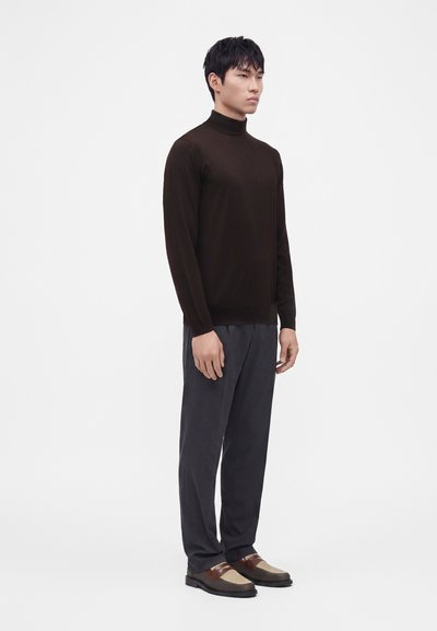 Homme adulte debout portant un col roulé marron foncé, un pantalon ample gris foncé et des chaussures habillées tissées marron et beige sur un fond blanc uni.