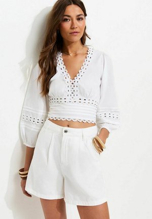 Donna che indossa una camicetta bianca corta con dettagli in pizzo e pantaloncini bianchi a vita alta, accessoriata con braccialetti beige e una collana.