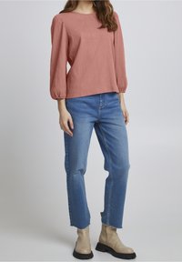 Maglione lungo a maniche lunghe di colore ruggine, testo con jeans chiari a vita alta e stivaletti beige con suola nera.