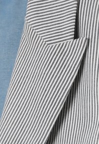 Gros plan d'un blazer blanc avec de fines rayures verticales noires sur un fond en tissu bleu texturé.