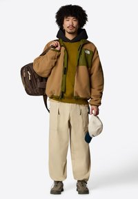 Giacca in pile color beige e verde, indossata sopra un felpa verde, abbinata a pantaloni cargo beige. Zaino marrone e cappellino crema tenuto in mano.
