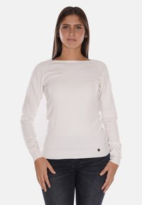 Maglione bianco a maglia con maniche lunghe, collo a barca, orlo a coste e un piccolo logo in basso a sinistra. Abbinato a jeans in denim scuri.