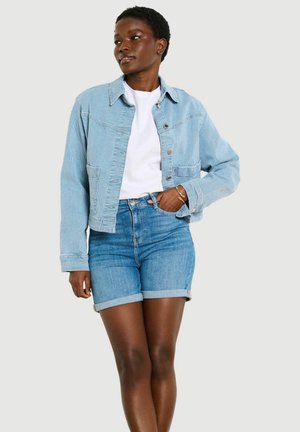 Kobieta w jasnoniebieskiej kurtce sztruksowej na białej koszuli i podwiniętych dżinsowych spodenkach, stojąca z jedną ręką w kieszeni na jednolitym tle.