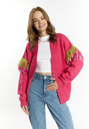 Roze gebreide cardigan met groene en roze franjes op de mouwen, gecombineerd met een witte top en high-waisted blauwe spijkerbroeken. Casual pasvorm.