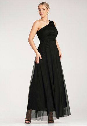 Schwarzes Maxikleid mit einem Schulterträger, plissiertem Oberteil, fließendem Tüllrock und seitlichem Schlitz. Das transparente Gewebe erzeugt einen geschichteten Effekt. Offene High Heels.