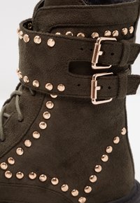 Bottines en daim vert olive avec des sangles à double boucle, des rivets en bronze sur le côté et une surface texturée pour plus de détails.