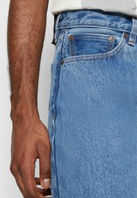 Gros plan sur le bras et la main d'une personne à côté d'un jean en denim bleu clair avec les poches avant et la poche à monnaie visibles.