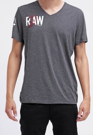 Personne portant un T-shirt à col en V gris foncé avec l'inscription « RAW » en blanc et rouge sur la poitrine gauche, et un pantalon noir, debout les bras détendus.