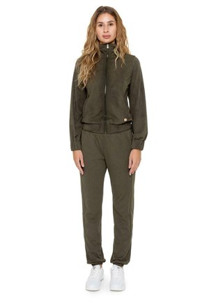 Une femme porte une veste zippée vert olive foncé et un pantalon jogger assorti, avec des baskets blanches, sur un fond blanc uni.