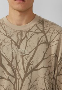Il felpa beige presenta un motivo di rami d'albero in marrone scuro, con il nome del brand "SOLIVER" e "ELEVATED SINCE 1969" stampati sul davanti.