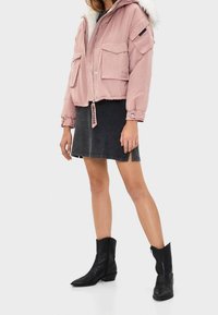 Rosa cropped jakke med frontlommer og pelsforet hætte, parret med en sort denimnederdel og sorte cowboystøvler. Glat tekstur, moderne design.