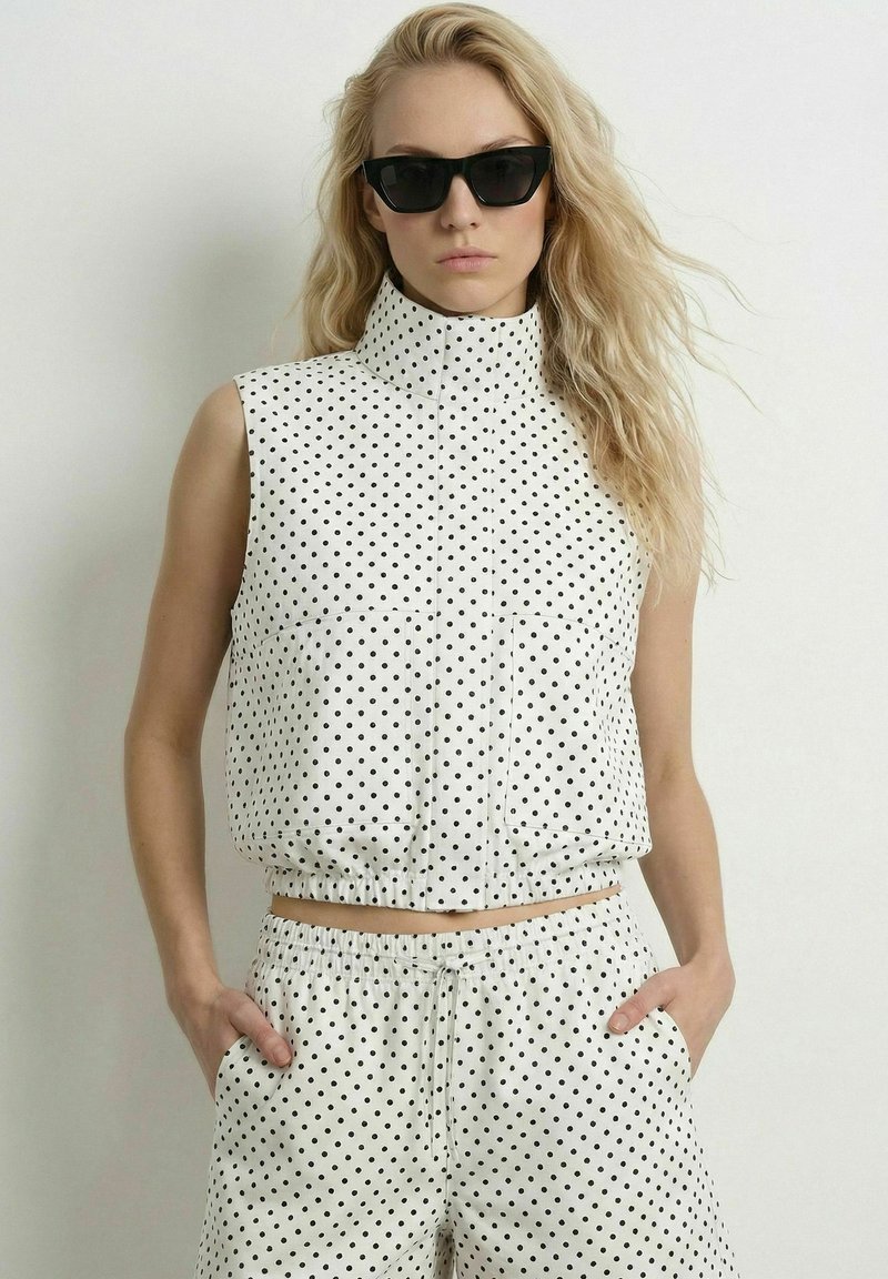 Femme portant des lunettes de soleil noires et un haut blanc sans manches à pois avec un pantalon assorti à pois, debout les mains dans les poches.