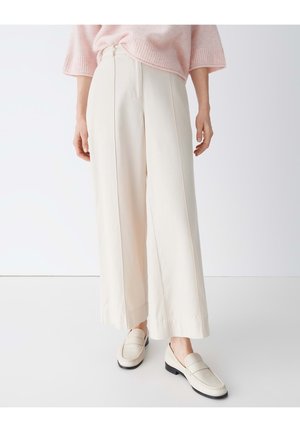 Frau trägt cremefarbene Hosen mit weitem Bein, cremefarbene Loafers und einen hellrosa Pullover, steht vor einem schlichten weißen Hintergrund.