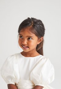 Robe blanche avec des manches bouffantes, texture lisse, encolure ronde et taille ajustée. L'enfant a les cheveux noirs et un léger sourire.